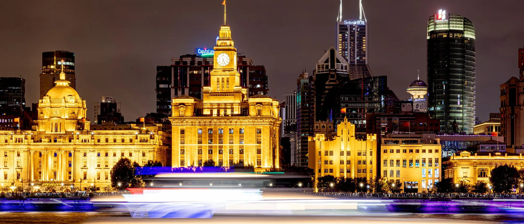 The Bund's historic buildings glowing gold on the Huangpu's west bank at night, with a clock tower and a boat's light trails streaking across the water